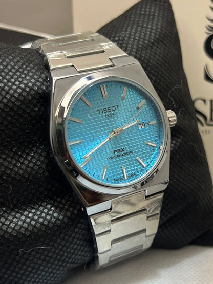 A Tissot PRX Powermatic watch with a bright blue dial and a silver stainless steel case and bracelet, displayed on a black textured cushion with a branded watch box in the background.